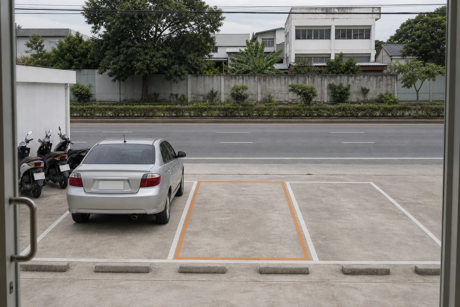 Seen from the shop door: the pale grey concrete apron with three white-marked bays, an ordinary silver sedan in the left one, the middle one outlined in soft orange as the pickup bay, motorbikes along the left wall, and the four-lane road beyond