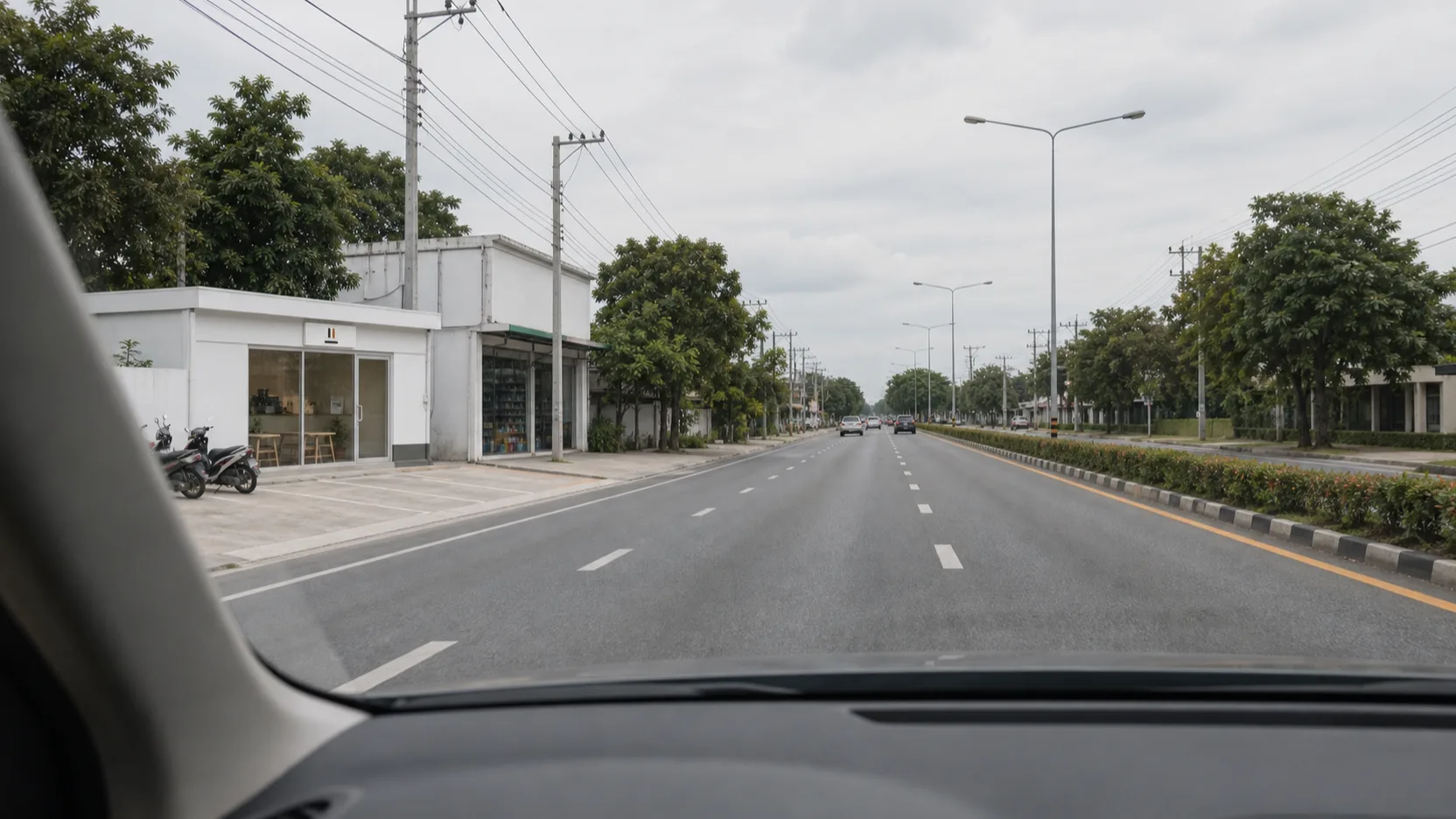 A driver's-eye view through the windscreen along a four-lane road with a planted median, the shop's white unit and small white signboard ahead on the left, with the pharmacy just beyond it
